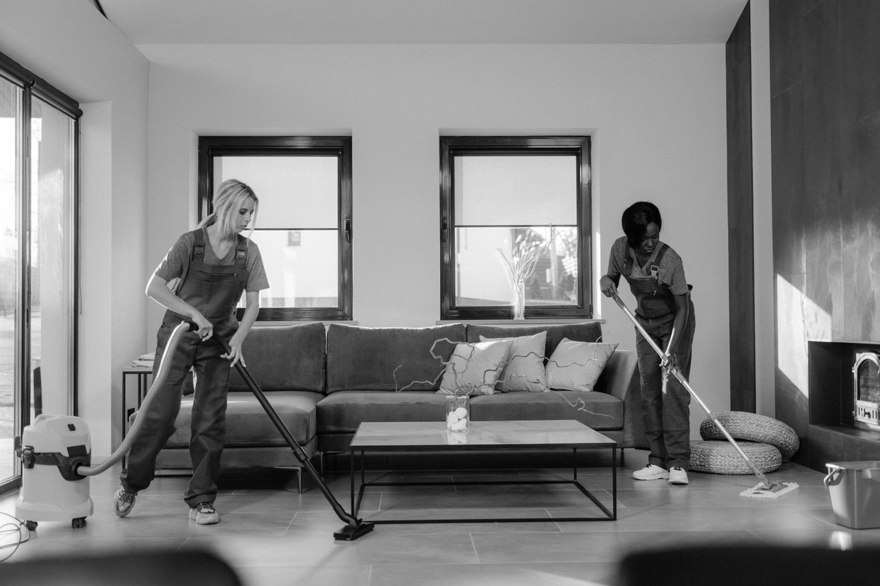 Mastering the First Impression: Your intriguing post title goes here Two women in work clothes cleaning a modern living room using a vacuum and mop.